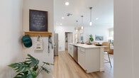This well-designed single-floor residence offers a bright, contemporary living experience with a cohesive aesthetic carried throughout every room. The open-concept kitchen serves as the heart of the home, showcasing a large center island with quartz countertops, a stainless steel sink, and pendant lighting overhead. White shaker-style cabinetry lines the walls alongside a full suite of stainless steel appliances, including a refrigerator, microwave, and range. Light wood-look flooring flows seamlessly from the kitchen into the adjoining dining and living areas, creating a unified, airy feel. The living room is bathed in natural light from large windows fitted with blinds, and features elevated ceilings that add to the sense of openness. The primary bedroom is generously sized with plush carpeting, a ceiling fan, and direct access to an en suite bathroom complete with a double-sink vanity topped with quartz countertops and a walk-in shower. A spacious walk-in closet with dual hanging rods provides ample storage. A secondary bedroom also features carpet flooring, a bright window, and its own walk-in closet. The secondary bathroom includes a single-sink vanity with quartz countertops and a full tub-shower combination. Throughout, the clean white palette and modern fixtures create a fresh, inviting ambiance.