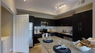 This single-floor space showcases a thoughtfully designed interior that balances modern aesthetics with practical functionality. The kitchen serves as the centerpiece of the space, featuring rich, dark cabinetry that provides a bold contrast against the light granite countertops. A spacious kitchen island offers additional prep and dining space, comfortably seating multiple guests. The full suite of stainless steel appliances — including a refrigerator, electric range, built-in microwave, and dishwasher — lends a polished, contemporary feel. A classic white subway tile backsplash adds texture and visual interest, complementing the overall design palette. Track lighting overhead ensures the kitchen is well-lit and functional, while the neutral tile flooring ties the space together seamlessly. The layout is open and efficient, with ample cabinetry offering generous storage throughout. The overall ambiance is modern and refined, with clean lines, upscale finishes, and a cohesive color scheme that creates an inviting yet sophisticated atmosphere suited for both everyday living and entertaining.