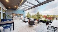 Located at 633B SW 150th St in Burien, Washington, this contemporary rooftop terrace is a thoughtfully designed outdoor amenity space that seamlessly blends sophisticated style with functional outdoor living. The terrace is finished with large-format porcelain tile pavers throughout, providing a clean, cohesive foundation for the entire space.

A standout architectural feature is the pergola structure spanning a portion of the terrace, constructed with dark-stained wood beams and a translucent polycarbonate roof that allows natural light to filter through while providing shelter from the elements. Recessed lighting integrated into the pergola ensures the space remains usable into the evening hours.

The outdoor kitchen area is fully equipped with a stainless steel gas grill, stone tile countertops, and custom wood-accented cabinetry — ideal for entertaining. Adjacent to the kitchen, multiple seating configurations are arranged throughout the terrace, including lounge chairs, chaise lounges, and accent tables with decorative planters adding natural greenery.

A dedicated dining area features dark metal tables paired with vibrant blue chairs, positioned near floor-to-ceiling glass walls that connect the terrace to the interior. The open railing along the terrace perimeter frames panoramic views of the lush Pacific Northwest tree canopy and seasonal foliage, creating a serene, elevated outdoor retreat.
