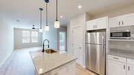 Located at 4408 N Church St in Greensboro, NC, this well-appointed one-bedroom, one-bathroom unit offers a thoughtfully designed single-floor layout with a clean, modern aesthetic throughout. The open-concept kitchen and living area serves as the heart of the home, anchored by a central island with granite countertops, a stainless steel sink, and stylish pendant lighting. The kitchen is fully equipped with white shaker cabinets, granite countertops, stainless steel appliances including a refrigerator, electric range, and built-in microwave, plus a wall-mounted range hood — all set atop warm, light-toned hardwood-style flooring that flows seamlessly through the main living areas. The living room provides a bright, open canvas with neutral walls and continued hardwood-style flooring, ideal for a variety of furniture arrangements. The bedroom is comfortably carpeted, features two windows that invite natural light and outdoor views, and is equipped with a ceiling fan. The full bathroom offers hardwood-style flooring, a granite-topped vanity with white cabinetry, and a shower-tub combo. A dedicated laundry room with washer/dryer hookups and built-in wire shelving adds everyday convenience. The unit presents a fresh, move-in-ready feel with a cohesive neutral palette throughout.