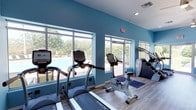 This single-floor fitness center is designed to provide an energizing and comfortable workout environment. The space is instantly recognizable by its bold teal-painted walls, which create a vibrant and motivating atmosphere throughout. Large, floor-to-ceiling windows line one wall, flooding the room with abundant natural light and offering unobstructed views of an outdoor pool area and surrounding green landscaping — allowing exercisers to enjoy a refreshing outdoor connection while working out indoors.

The gym is outfitted with a comprehensive selection of cardio equipment from the True Fitness brand, including multiple treadmills, an upright stationary bike, and a stair climber machine, all arranged to maximize the natural light and views. The light wood-toned flooring adds warmth to the space and complements the bright, airy aesthetic.

Practical features include recessed and flush-mounted ceiling lighting to ensure the space remains well-lit at all hours, along with clearly marked emergency exit signage and HVAC vents for climate control. The overall layout is open and spacious, providing ample room to move between machines comfortably. This fitness center is a thoughtfully designed amenity space that balances functionality with an inviting, light-filled ambiance.