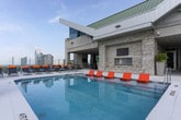Located at 3390 Stratford Rd NE in Atlanta's prestigious Buckhead district, this amenity floor delivers a sophisticated urban retreat with a striking blend of modern design and high-end functionality. The crown jewel of the space is a resort-style rooftop pool framed by vibrant orange and navy lounge chairs, offering sweeping panoramic views of the Atlanta skyline — an ideal setting for relaxation above the city. Adjacent to the pool, a spacious lounge area features an open, flowing layout with tray ceilings, floor-to-ceiling windows, and abundant natural light. Deep-toned hardwood-style flooring, plush blue velvet accent chairs, neutral ottomans, and rich chocolate sofas create a refined yet comfortable atmosphere. Pendant lighting in warm amber tones adds visual interest over a dining or social gathering area, further enhancing the inviting ambiance. A dedicated conference room provides a more private setting, outfitted with a large dark wood table, upholstered seating, a wall-mounted display, and a contemporary fireplace — all framed by city views through expansive glass. The entry corridor features bold blue chevron-patterned wallcovering, tile flooring, and digital information displays, setting an energetic and polished tone from arrival.