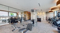 Located at 15 Artisan in Salem, New Hampshire, this well-appointed fitness center offers a comprehensive workout environment within a single, open-plan floor. The space is designed with a clean, modern aesthetic, featuring neutral-toned carpet flooring, crisp white walls, and a striking geometric accent wall that adds a bold visual focal point to the exercise area. Warm pendant lighting with large drum shades complements the abundant natural light that pours in through expansive floor-to-ceiling windows, offering open views of the surrounding exterior. The cardio zone is equipped with treadmills, ellipticals, and stationary cycling bikes positioned to take full advantage of the outdoor views, while wall-mounted flat-screen TVs provide additional entertainment. The strength training area features a multi-functional cable machine, free weights, benches, and dedicated storage shelving for equipment organization. A TRX suspension training station and medicine balls round out the functional fitness offerings. Large mirrors along one wall visually expand the space and support proper form during workouts. A glass-paneled entryway with wood-accented framing provides a polished, welcoming entrance. The overall ambiance is bright, energetic, and thoughtfully designed to support a wide range of fitness activities.