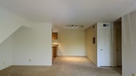 This single-floor space in Brandywine presents an open and functional layout with a straightforward design. The main living area is finished with wall-to-wall carpeting in a neutral tone, contributing to a clean and understated aesthetic. A textured ceiling adds subtle visual interest overhead, while a recessed tray ceiling detail in the central area of the room is highlighted by a track lighting fixture, providing focused illumination across the space. The walls are painted in a light cream tone, lending a bright and airy feel to the interior. A partial view into the kitchen reveals warm wood-toned cabinetry, an upper cabinet, and a countertop with a sink, suggesting a compact but practical cooking space. A wall cutout connects the kitchen to the living area, enhancing the sense of openness between the two spaces. A standard interior door on one side of the living area provides access to an additional room or closet. Ventilation registers are visible along the walls and ceiling, indicating a centralized HVAC system. The overall layout is simple and versatile, offering a functional foundation suitable for a variety of furnishing arrangements.