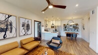 This contemporary single-floor residence showcases a thoughtfully designed open-concept layout that seamlessly connects the living, dining, and kitchen areas. The living room is anchored by a stylish wood-blade ceiling fan, recessed lighting, and rich hardwood-style flooring, complemented by a textured shag area rug and modern furnishings in warm cognac leather and sleek black accents. A sliding glass door provides access to an outdoor balcony, bringing in ample natural light. The kitchen features white shaker cabinetry, stainless steel appliances including a side-by-side refrigerator and range with a wall-mounted ventilation hood, white subway tile backsplash, and a central island with light countertops. Decorative open wood shelving adds a functional design element to the dining area. The bedroom offers a tray ceiling, ceiling fan, a walk-in closet, and direct views into the living space, creating a connected yet private retreat. Two bathrooms are finished with marble-style countertops, backlit vanity mirrors, floating white cabinetry, large-format tile flooring, and frameless glass shower enclosures, delivering a clean and refined aesthetic throughout.