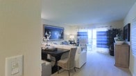 This single-floor space presents a thoughtfully designed open-concept layout that integrates the dining and living areas into one cohesive, inviting environment. The interior carries a distinct coastal aesthetic, highlighted by a rich navy and white color palette that runs throughout the furnishings and decor. A striking large-format artwork depicting sailboats serves as a focal point on the wall, lending a nautical charm to the space.

The dining area features a dark wood table paired with a mix of woven and upholstered seating, creating an elegant yet relaxed atmosphere. It flows seamlessly into the living room, which is furnished with plush white sofas accented by blue throw pillows, reinforcing the space's cohesive design theme.

Soft, neutral-toned carpeting extends throughout, adding warmth and comfort underfoot. The living area is bathed in natural light, which streams in through a glass sliding door framed by bold blue and white striped curtains. The door opens to an outdoor balcony, providing a bright and airy connection to the exterior.

Overhead, smooth ceilings and well-placed lamps contribute to a warm, ambient lighting scheme that complements the natural light. Overall, the space offers a clean, well-coordinated interior with a relaxed coastal sensibility.