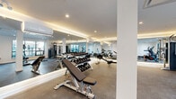 Located at 750 Robinson Street in Coquitlam, BC, this modern fitness facility offers a thoughtfully designed exercise space on a single floor. The room features a clean, contemporary aesthetic with crisp white walls, recessed pot lighting, and warm accent lighting along the ceiling perimeter that creates an inviting atmosphere.

The exercise room is well-appointed with a diverse selection of equipment, including a treadmill, elliptical trainer, rowing machine, stationary bike, and a multi-station cable machine for strength training. A full dumbbell rack paired with an adjustable weight bench provides ample options for free weight workouts. Floor-to-ceiling mirrors line one wall, enhancing the sense of space and allowing users to monitor their form during exercise. Rubber flooring throughout provides safety and durability underfoot.

Natural light filters in through large windows along one wall, offering views of the surrounding neighbourhood and keeping the space feeling bright and open. A wall-mounted air conditioning unit ensures comfortable year-round use. A built-in water bottle refilling station is conveniently located near the entrance corridor, which features a sleek glass-and-black-frame door. The overall design is functional yet polished, delivering a professional gym experience in a well-maintained, accessible setting.