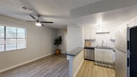 This single-floor residence offers a clean, modern aesthetic with a cohesive neutral color palette and wood-style flooring that runs throughout the entire space. The open-concept layout seamlessly connects the kitchen and living area, creating a functional and inviting environment. The kitchen is well-equipped with white shaker-style cabinetry, a classic white subway tile backsplash with an accent tile detail, dark countertops, stainless steel appliances including a dishwasher and full-size refrigerator, and a peninsula counter that serves as a natural divider between the kitchen and living space. The living area is bright and airy, featuring a ceiling fan with light and a large window that allows natural light to filter in. A dedicated entry area with a paneled front door and sidelight window provides a welcoming arrival experience. The bedroom is generously sized, offering a sliding closet, a ceiling fan, and a window with outdoor views. Textured ceilings are present throughout. The overall ambiance is fresh and move-in ready, reflecting a straightforward, practical design with contemporary touches.
