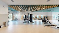 This expansive fitness center in Clermont, FL offers a thoughtfully designed exercise space spread across a single floor. The layout is divided into distinct workout zones, providing ample room for a comprehensive range of training activities. Architecturally, the space stands out with its soaring ceilings accented by striking wooden slat details set against a bold teal ceiling panel, creating a modern and energizing atmosphere. A ceiling fan and recessed lighting ensure comfort throughout the facility.

The cardio zone features multiple treadmills, stair climbers, and stationary bikes, several equipped with integrated digital screens. The strength training area includes a multi-functional cable machine, a dumbbell rack with accompanying bench, and full-length mirrors that enhance the sense of space. A dedicated functional training zone offers open floor space with a large tire, medicine balls, resistance equipment, and a wall-mounted storage rack for accessories.

Natural light pours in through multiple windows along the perimeter, offering outdoor views of the surrounding greenery. A glass roll-up garage door in the functional training area opens directly to the outdoors, allowing for fresh air and expanded workout possibilities. The flooring transitions between terrazzo, rubberized, and carpet tile surfaces to suit each training zone, completing this well-appointed and versatile fitness facility.