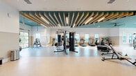 This expansive fitness center in Clermont, FL offers a thoughtfully designed exercise space spread across a single floor. The layout is divided into distinct workout zones, providing ample room for a comprehensive range of training activities. Architecturally, the space stands out with its soaring ceilings accented by striking wooden slat details set against a bold teal ceiling panel, creating a modern and energizing atmosphere. A ceiling fan and recessed lighting ensure comfort throughout the facility.

The cardio zone features multiple treadmills, stair climbers, and stationary bikes, several equipped with integrated digital screens. The strength training area includes a multi-functional cable machine, a dumbbell rack with accompanying bench, and full-length mirrors that enhance the sense of space. A dedicated functional training zone offers open floor space with a large tire, medicine balls, resistance equipment, and a wall-mounted storage rack for accessories.

Natural light pours in through multiple windows along the perimeter, offering outdoor views of the surrounding greenery. A glass roll-up garage door in the functional training area opens directly to the outdoors, allowing for fresh air and expanded workout possibilities. The flooring transitions between terrazzo, rubberized, and carpet tile surfaces to suit each training zone, completing this well-appointed and versatile fitness facility.