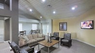 This single-story space offers a welcoming and functional layout centered around a comfortable living area. The room is defined by its distinctive wood-planked ceiling, which adds a rustic yet refined character to the space. Recessed lighting is strategically placed throughout, providing warm, even illumination across the room.

The living area is generously sized, accommodating a plush, cushioned sofa adorned with decorative pillows, alongside two leather accent chairs that add a touch of classic sophistication. A dark wood coffee table serves as the focal point of the seating arrangement, styled with decorative candle holders that contribute to the room's warm ambiance.

French doors with glass panes invite natural light into the space while providing access to the exterior, enhancing the open feel of the room. A framed wall mirror adds depth and visual interest to the neutral-toned walls.

Adjacent to the living area, a dining space is visible, furnished with a wooden table and chairs, making the layout practical for everyday living. Neutral carpet and light-toned walls throughout create a cohesive and inviting atmosphere well-suited for comfortable daily use.