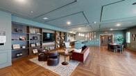 Located at 4733 S Packard Ave in Cudahy, Wisconsin, this single-floor community amenity space offers a thoughtfully designed combination of a kitchen and living room. The interior showcases striking herringbone-patterned hardwood flooring throughout, complemented by teal-toned walls with elegant wainscoting and coffered ceiling details that lend architectural character to the space.

The kitchen area is outfitted with rich walnut cabinetry, a quartz countertop with a full marble-look backsplash, a matte black sink and faucet, and stainless steel appliances. A bold teal island with white countertop seating for multiple bar-stool guests serves as the centerpiece, illuminated by a trio of pendant lights. A floating wood shelf above the counters is accented by botanical-patterned wallpaper, adding a vibrant design element.

The adjoining living area provides a warm, lounge-like atmosphere with curated furniture groupings, framed wall art, and built-in shelving. Large windows and glass sliding doors flood the space with natural light and connect directly to a generous outdoor terrace. The patio features ample seating, dining tables, a gas grill, and open views of the surrounding multi-story building and neighborhood skyline, creating an inviting setting for gathering and relaxation.
