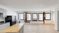 Located at 511 Maple Blvd in Kansas City, MO, Unit 6 is a thoughtfully laid-out single-floor unit that makes efficient use of its space. The open main living area is finished with light wood-look flooring throughout, lending the space a clean and modern feel. A row of large windows with bold black frames lines one wall, allowing natural light to pour in while offering views of the surrounding neighborhood. A ceiling fan provides additional comfort, and an in-unit washer is conveniently tucked along the wall. A charming exposed brick column adds a touch of character and industrial heritage to the otherwise contemporary interior. The kitchen is well-appointed with gray cabinetry, dark countertops, and a full suite of black appliances including a gas range, dishwasher, microwave, and refrigerator. A freestanding island with a butcher block top provides additional prep and storage space. Recessed lighting illuminates the kitchen area, while a hallway connects to the bathroom and entry door, keeping the layout practical and organized. The white walls and neutral palette throughout create a bright, versatile environment. This unit at 511 Maple Blvd offers a functional and stylish living space in Kansas City's 64124 zip code.
