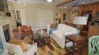 This single-floor home offers a comfortable and well-appointed living space with a warm, welcoming ambiance. The living room serves as the heart of the home, featuring warm beige walls complemented by an exposed wooden beam that adds a rustic architectural touch. Crown molding along the ceiling enhances the classic character of the space.

The room is furnished with a slipcovered white sofa adorned with colorful decorative pillows and a cozy teal throw blanket, paired with a rich cognac leather armchair. A decorative metal coffee table with an ornate base sits atop a multicolored area rug, layered over hardwood flooring that runs throughout. A traditional wooden side table with turned legs adds further charm.

A set of double doors with a classic paneled design provides closet or storage access, while an open passageway leads to adjacent areas, including what appears to be a kitchen space with hanging cookware visible. The layout flows naturally between rooms, creating a sense of openness. Ceiling fans with light fixtures provide practical comfort, while the overall design aesthetic blends traditional and eclectic elements harmoniously.