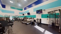 This fitness center is a fully equipped, single-floor facility designed to support a comprehensive workout experience. The space is immediately distinguished by its bold, color-blocked wall design featuring horizontal stripes of navy blue, teal, and white, which lend the room a vibrant and energetic atmosphere. The ceilings rise generously overhead, contributing to an open and airy feel, while ceiling fans and recessed lighting ensure the space remains well-ventilated and brightly lit throughout.

The flooring consists of durable rubber gym matting, providing both safety and comfort underfoot. Cardio equipment lines one side of the room and includes multiple treadmills, stair climbers, and a recumbent bike, offering a range of options for cardiovascular training. Wall-mounted flat-screen TVs are positioned above the cardio stations, allowing users to stay entertained during workouts.

The strength training area is equally well-appointed, featuring a variety of cable machines, a leg press, a lat pulldown station, free weights, benches, and medicine balls. A large mirror spans one wall, enhancing the sense of space and allowing users to monitor their form. Fitness center rules are posted near the entrance, and natural light filters in through a glass door. The overall environment is clean, organized, and purpose-built for effective fitness training.