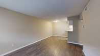 Located at 10925 Beamer Rd in Houston, TX 77089, this single-floor one-bedroom unit offers a practical and well-organized layout with consistent dark hardwood-style flooring throughout. The living room serves as the central gathering space, characterized by warm neutral-toned walls, clean white baseboards, and a large window fitted with horizontal blinds that allows natural light to fill the room. A front entry door is positioned adjacent to the window, providing direct access to the space. The kitchen and living room share an open-concept arrangement, with the kitchen defined by a raised peninsula counter that separates the two areas. The kitchen features white cabinetry, a countertop with a built-in sink, and white appliances, including a range and refrigerator, all set beneath under-cabinet lighting. A hallway connects the main living area to the private bedroom, which is generously sized and equipped with a ceiling fan with light fixture. The bedroom benefits from natural light via a window with blinds and includes a dedicated closet with a hanging rod for organized storage. The overall ambiance of the unit is clean, neutral, and move-in ready, with a cohesive color palette of beige and white tones that lends a bright, understated character throughout.