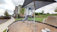 This single-floor residence is part of a well-maintained multi-building community set on gently rolling, landscaped grounds. The unit itself includes a spacious walk-in closet finished with wall-mounted wire shelving for organized storage, soft carpeted flooring, and neutral-toned walls that create a clean, move-in-ready atmosphere. A dedicated laundry area is equipped with hookups, offering added convenience within the unit.

The community grounds are a notable feature of this property, offering generous green space and two separate outdoor play areas. The first playground includes a shaded metal slide structure positioned over a wood-chip safety surface, sheltered by a large canopy overhead. The second play area features a dome-style rope climbing structure with stepping platforms, set against a backdrop of open lawn and a columned community pavilion. Both areas are well-suited for active outdoor use and are situated amid neatly landscaped surroundings with young trees and maintained turf. The overall environment reflects a thoughtfully designed residential community with practical interior features and engaging shared outdoor spaces.