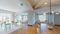 This single-story space presents a well-designed, contemporary layout that serves multiple functional purposes. Polished amber-toned concrete floors run throughout, providing a warm, cohesive foundation that contrasts with the cool gray walls and white ceilings accented by exposed beam detailing.

A glass-walled conference room is a standout feature, offering transparency and accessibility while accommodating a full conference table and ergonomic rolling chairs—ideal for meetings or collaborative work. Adjacent to this, an open lounge area features a plush sectional sofa, leather accent chairs, a round coffee table, and framed artwork, creating a relaxed yet refined atmosphere. Large sliding glass doors and floor-to-ceiling windows flood the space with abundant natural light and provide views of surrounding greenery.

A dedicated kitchenette area is equipped with white cabinetry, a Keurig coffee station, a beverage refrigerator, a printer, and a wall-mounted television backed by a striking geometric-patterned accent wall. High-top bistro seating near the windows offers an additional casual gathering spot. The overall ambiance is modern, clean, and inviting, balancing professional functionality with comfortable, communal living.