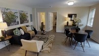 This single-floor residence offers a thoughtfully designed layout that blends comfort and functionality. The open-concept living and dining area serves as the heart of the home, featuring smooth light-toned hardwood flooring that runs throughout, creating a cohesive and inviting atmosphere. Neutral wall tones in soft white and warm beige provide a clean backdrop that enhances the sense of brightness and openness.

The living area is anchored by a classic sofa styled with accent pillows and a cozy throw, complemented by a mid-century modern armchair and a sleek geometric coffee table set atop a patterned area rug. Large framed wall art adds a bold decorative touch to the space. Adjacent to the living area, the dining space accommodates a round wooden table with contemporary chairs, accented by a stylish table lamp and floral arrangement that bring warmth to the corner.

Two doorways visible from the main living area provide access to additional rooms, including what appears to be a bathroom and a bedroom, suggesting a well-connected and functional floor plan. Flush-mount ceiling lighting ensures consistent illumination throughout. Overall, this residence presents a modern, well-balanced interior that is both practical and aesthetically refined.