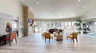 This single-story clubhouse in Vacaville, California, offers a generously sized, open-concept layout designed to serve as a multifunctional community gathering space. The interior is anchored by soaring vaulted ceilings that amplify the sense of openness throughout. Light gray wood-style flooring runs continuously across the space, unifying each functional area seamlessly.

The main lounge area is a standout feature, centered around a dramatic floor-to-ceiling stacked stone fireplace with an inset flat-screen television. A curved wall of large windows and glass doors floods the lounge with abundant natural light and provides unobstructed views of the outdoor pool and patio area. Comfortable seating arrangements, including a leather sofa, tufted bench, and accent chairs, create an inviting atmosphere.

Adjacent to the lounge, a kitchenette is outfitted with granite countertops, dark cabinetry, a stainless steel dishwasher, and a coffee station. A dining area with bar-height seating and a formal desk workspace are also present, along with a dedicated fitness center enclosed by glass-panel French doors, equipped with cardio machines. A private office area completes the layout, offering a functional workspace with natural light.