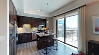 This single-floor residence showcases a thoughtfully designed open-concept layout that seamlessly blends the kitchen and living areas into one inviting space. The kitchen is a standout feature, appointed with rich dark espresso cabinetry that contrasts beautifully against light granite countertops and a classic tile backsplash. A central island with bar seating provides an ideal space for casual dining or entertaining. Stainless steel appliances, including a gas range, dishwasher, and range hood, complete the well-equipped culinary space. Track lighting and elegant pendant fixtures illuminate the kitchen, enhancing the warm, contemporary atmosphere. Wide-plank hardwood flooring flows throughout, adding warmth and continuity to the design. Floor-to-ceiling sliding glass doors draw the eye toward a spectacular panoramic city view, flooding the interior with abundant natural light. The doors open directly onto a private balcony, offering an elevated outdoor vantage point overlooking the urban skyline and surrounding cityscape. The overall ambiance of the space is modern and sophisticated, combining high-end finishes with functional design to create a residence that is both stylish and comfortable.