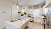 This single-story home is designed with a clean, contemporary aesthetic, combining functionality and style throughout its thoughtfully arranged spaces. The kitchen serves as a central feature, showcasing white shaker-style cabinetry, a white quartz countertop island with bar seating, and a full suite of stainless steel appliances including a refrigerator, dishwasher, and microwave. Glass pendant lights hang above the island, complementing recessed ceiling lighting, while wood-look flooring and multiple windows create a bright, welcoming atmosphere. A subway tile backsplash adds a refined detail to the overall design.

The living area is open and well-lit, featuring large sliding glass doors that provide direct access to a private outdoor space enclosed by lush greenery. A ceiling fan, recessed lighting, and generous window placement ensure the room remains comfortable and airy throughout the day.

The bedroom is a serene retreat, featuring plush carpeting, ample natural light from a well-positioned window, and a ceiling fan overhead. The ensuite bathroom offers a glass-enclosed walk-in shower with large-format tile surround, a white vanity with stone-topped counters, wood-look flooring, and direct access to a walk-in closet, completing this comfortable and cohesive living space.