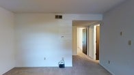 This single-floor residence offers a simple, functional layout designed for comfortable everyday living. The main living area is finished with wall-to-wall carpeting in a neutral tone, contributing to a quiet and understated atmosphere throughout. Crisp white walls provide a clean, bright backdrop, while a ceiling-mounted HVAC vent ensures climate control throughout the space. A thermostat is conveniently positioned on the wall, reflecting the unit's practical appointments. From the living area, a hallway extends toward the private quarters of the home, with doorways leading to additional rooms — including what appears to be a bedroom with a dark-toned door, adding a subtle contrast to the otherwise light interior. A second doorway is also visible along the corridor, suggesting multiple rooms accessible from the central hall. The overall design is straightforward and efficient, with a neutral color palette of whites and beiges that lends itself well to personalization. The space is move-in ready and offers a blank canvas for a variety of furnishing styles. Recessed lighting and consistent flooring materials tie the rooms together, creating a cohesive and functional living environment.