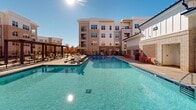 Located at 15101 Royal Creek Dr in Fuquay-Varina, NC, this generously sized outdoor amenity space is thoughtfully designed for communal living and recreation. The centerpiece is a large, resort-style swimming pool with crystal-clear blue water, surrounded by lounge chairs, colorful umbrellas, and a dedicated pool deck that invites relaxation under the open sky. Adjacent to the pool, a spacious concrete courtyard serves as a social hub, furnished with vibrant yellow chairs and dark bistro tables arranged for casual gathering. Overhead string lights crisscross between tall poles, creating an inviting ambiance for evening use. A built-in outdoor grilling station provides a convenient space for outdoor cooking and entertaining. Thoughtfully landscaped planters filled with river rock and ornamental plantings add texture and visual interest throughout the area, softening the hardscape with natural elements. A lush green lawn area with swing seating offers a quieter retreat within the broader outdoor environment. The multi-story residential building surrounds the courtyard on multiple sides, constructed with a modern combination of brick, fiber cement siding, and large windows. Wide, accessible walkways connect each zone of the space seamlessly, making this outdoor area a well-rounded and functional amenity for its community.