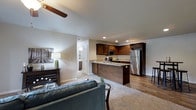 This single-floor space is thoughtfully designed with an open-concept layout that seamlessly connects the living area and kitchen, creating a cohesive and functional environment. The kitchen is a standout feature, showcasing rich dark-stained cabinetry that extends to the upper walls, offering ample storage. Light-toned granite or stone countertops provide a striking contrast to the cabinetry, while stainless steel appliances — including a refrigerator and built-in microwave — lend a contemporary, polished feel. A peninsula with a built-in sink serves as both a practical workspace and a natural divider between the kitchen and living areas.

The living space is carpeted and anchored by a plush area rug, creating a warm and inviting atmosphere. A ceiling fan overhead ensures comfortable airflow throughout. Neutral grey walls and recessed lighting contribute to a clean, modern aesthetic. Hardwood-style flooring in the kitchen and transition areas adds visual depth and durability. A hallway visible from the main area hints at additional rooms beyond, including what appears to be a bathroom with white cabinetry. Overall, the space balances practicality with contemporary style.