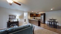 This single-floor space is thoughtfully designed with an open-concept layout that seamlessly connects the living area and kitchen, creating a cohesive and functional environment. The kitchen is a standout feature, showcasing rich dark-stained cabinetry that extends to the upper walls, offering ample storage. Light-toned granite or stone countertops provide a striking contrast to the cabinetry, while stainless steel appliances — including a refrigerator and built-in microwave — lend a contemporary, polished feel. A peninsula with a built-in sink serves as both a practical workspace and a natural divider between the kitchen and living areas.

The living space is carpeted and anchored by a plush area rug, creating a warm and inviting atmosphere. A ceiling fan overhead ensures comfortable airflow throughout. Neutral grey walls and recessed lighting contribute to a clean, modern aesthetic. Hardwood-style flooring in the kitchen and transition areas adds visual depth and durability. A hallway visible from the main area hints at additional rooms beyond, including what appears to be a bathroom with white cabinetry. Overall, the space balances practicality with contemporary style.