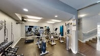Located at Teal Landing in O'Fallon, Missouri, this fitness center is a comprehensively equipped exercise facility spread across a single floor. The space is divided into two functional zones designed to support a full range of workout routines.

The main cardio and strength room features rubber flooring and is lined with large wall-to-wall mirrors that enhance the sense of space and allow exercisers to monitor their form. The room is outfitted with a selection of True-brand cardio equipment, including treadmills, elliptical trainers, upright bikes, and cycling bikes. Overhead, bright flush-mount and recessed lighting ensures excellent visibility throughout.

A secondary room provides a dedicated space for free weights and functional training. A tiered dumbbell rack lines the wall alongside TRX suspension straps and resistance bands, offering versatile strength training options. This area also features stability balls, medicine balls, and a flat workout bench, making it ideal for stretching and full-body conditioning. Natural light filters in through multiple windows and a glass-panel exterior door, giving the room an open, airy feel with views to the outdoors.

The facility is accessed via full-height glass doors that open to an interior staircase, seamlessly connecting the fitness center to the broader Teal Landing community.