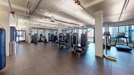 Located at 2025 Fulton Place in Reston, Virginia, this single-floor fitness center is a comprehensively outfitted facility designed to support a full range of workout routines. The space is organized into distinct functional zones, each tailored to a specific type of training. The main strength and machine area features a wide selection of selectorized cable and resistance machines arranged across durable rubber-tile flooring, complemented by an exposed ceiling with visible ductwork and pipeline infrastructure that lends the space an industrial, open feel. Large windows throughout the facility invite generous natural light and offer views of the surrounding outdoor environment. A dedicated cardio zone houses multiple treadmills, ellipticals, stair climbers, and rowing machines, all positioned to face outward-facing windows for an energizing workout experience. The free weights and functional training area is well-stocked with dumbbells, barbells, a punching bag, TRX suspension systems, and a variety of functional fitness accessories. Full-length mirrors line several walls, enhancing spatial openness and supporting proper form. A separate studio room, enclosed by glass-panel doors, features light hardwood-style flooring, a ballet barre, large circular pendant lights, a mounted display screen, and an array of accessories including stability balls — ideal for group fitness, yoga, or personal training sessions.