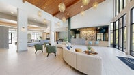This thoughtfully designed clubhouse in Jacksonville, FL delivers an upscale community experience across a single, expansive floor. The grand entrance greets visitors with soaring ceilings, floor-to-ceiling black-framed glass walls, and warm woven pendant lighting suspended from a rich wood-paneled ceiling, creating an immediate sense of openness and sophistication. Light-toned large-format tile flooring flows seamlessly throughout, unifying each distinct zone.

The central lobby features a welcoming reception desk accented by an eye-catching botanical wallcovering and complemented by a curved sectional sofa, earth-toned armchairs, and live greenery. A convenient refreshment station with built-in cabinetry and a mini-fridge offers residents easy access to everyday essentials.

Adjacent social spaces include a casual lounge with a tray ceiling and bold jewel-toned furnishings anchored by a statement chandelier, alongside a versatile gathering room with sliding wood panel doors, tufted leather coffee tables, and ample natural light. The social kitchen is a standout feature, boasting a generous stone-topped island with bar seating, a glass tile backsplash, open shelving, and warm pendant lighting.

A dedicated co-working area lined with expansive windows and a lush outdoor backdrop provides a bright, productive environment for residents seeking a professional workspace within the community.