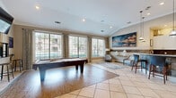 Located at 13100 W Bellfort Ave in Houston, TX, this well-appointed community amenity space combines recreation and comfort in a single, thoughtfully designed floor. The main lounge and game area is anchored by a full-size pool table set atop rich hardwood flooring, which transitions seamlessly into large-format diagonal tile throughout the rest of the space. A wall-mounted flat-screen TV and bar-height seating make this an ideal gathering spot for entertainment. Large windows dressed with patterned drapes flood the room with natural light while offering views of the outdoor pool area beyond. A vaulted ceiling accent and stone-clad wainscoting add architectural character, complemented by curated artwork and a cozy seating nook with upholstered accent chairs. The kitchenette is fully equipped with white shaker-style cabinetry, a stacked stone mosaic backsplash, light quartz countertops, stainless steel appliances including a side-by-side refrigerator and dishwasher, and a pendant-lit sink area. A separate bar-height communal table with metal stools provides additional casual seating. French doors with glass panels connect the spaces and enhance the open, airy feel of this inviting community hub.