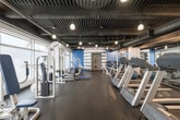 Located at 1431 Ocean Ave in Santa Monica, California, this fitness center offers a comprehensive workout environment designed to accommodate a full range of exercise needs. The single-floor space features an industrial-inspired aesthetic, with an exposed dark ceiling showcasing overhead ductwork, structural elements, and pendant lighting that casts a warm, focused glow throughout the facility.

The main floor is organized into distinct workout zones. A long row of treadmills and cardio machines lines one side of the space, providing ample options for cardiovascular training. Opposite these, a selection of cable machines and selectorized strength equipment — upholstered in blue and white — runs along floor-to-ceiling windows, allowing natural light to fill the space and offering views to the exterior.

A dedicated free weights area features a full rack of dumbbells alongside benches and additional strength machines, making it suitable for a variety of resistance training exercises. The rubber flooring throughout provides both safety and durability underfoot.

One of the most distinctive architectural elements is a cylindrical, horizontally striped gray and white structure housing an interior door with porthole-style accents — a visually striking design feature that adds a nautical character fitting of the seaside Santa Monica location. Wall-mounted mirrors further enhance the sense of openness across the facility.