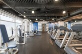Located at 1431 Ocean Ave in Santa Monica, California, this fitness center offers a comprehensive workout environment designed to accommodate a full range of exercise needs. The single-floor space features an industrial-inspired aesthetic, with an exposed dark ceiling showcasing overhead ductwork, structural elements, and pendant lighting that casts a warm, focused glow throughout the facility.

The main floor is organized into distinct workout zones. A long row of treadmills and cardio machines lines one side of the space, providing ample options for cardiovascular training. Opposite these, a selection of cable machines and selectorized strength equipment — upholstered in blue and white — runs along floor-to-ceiling windows, allowing natural light to fill the space and offering views to the exterior.

A dedicated free weights area features a full rack of dumbbells alongside benches and additional strength machines, making it suitable for a variety of resistance training exercises. The rubber flooring throughout provides both safety and durability underfoot.

One of the most distinctive architectural elements is a cylindrical, horizontally striped gray and white structure housing an interior door with porthole-style accents — a visually striking design feature that adds a nautical character fitting of the seaside Santa Monica location. Wall-mounted mirrors further enhance the sense of openness across the facility.