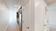 This single-floor unit showcases a clean, contemporary design throughout, with light wood-tone flooring and crisp white walls creating a bright and cohesive aesthetic. The kitchen is a functional highlight, featuring wood-grain cabinetry extending to the ceiling for ample storage, white quartz countertops, a subway tile backsplash, stainless steel appliances including a range and refrigerator, and a stainless undermount sink — all arranged in an efficient galley-style layout. A hallway connects the main living areas, with interior doors finished in white and fitted with brushed nickel hardware. The living area benefits from large windows that invite abundant natural light and provide views of the surrounding building exterior, while track lighting adds a practical and modern touch overhead. The bedrooms are well-proportioned, each featuring light flooring, white walls, and sliding closets with built-in hanging rods. One bedroom also enjoys its own window, enhancing the sense of brightness. The bathroom presents a minimalist design with a wall-mounted rectangular sink, a frameless mirror with integrated lighting, and tile flooring. Overall, the space offers a modern, move-in-ready environment with a neutral palette suited to a variety of interior styles.