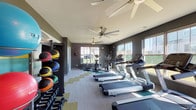 The fitness center at Villas of Jeffersonville is a thoughtfully designed single-floor facility that provides residents with a comprehensive workout environment. The interior features bold charcoal-gray walls accented by a striking color-block mural in teal, yellow, and white, lending the space a modern, energetic aesthetic. The flooring consists of patterned carpet tiles arranged in a dynamic chevron design incorporating teal, gold, and neutral tones, adding visual interest underfoot.

Natural light fills the room through numerous large windows lining multiple walls, offering views of the surrounding grounds and creating an open, airy atmosphere. Multiple ceiling fans are installed throughout the space, ensuring consistent airflow and comfort during workouts.

The cardio section is well-appointed with a row of treadmills positioned along the windowed walls, allowing exercisers to enjoy outdoor views while training. Elliptical machines and stationary bikes, including cycling-style bikes, round out the cardio offerings. A multi-station cable machine provides options for strength and resistance training, while a dedicated free weight and medicine ball rack along one wall supports functional fitness routines. A wall-mounted television adds entertainment options. A direct exterior door provides convenient access to the outdoors, completing this functional and visually engaging fitness space.