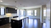 This single-floor space showcases a thoughtfully designed open-concept layout that seamlessly integrates the kitchen and living areas into one cohesive environment. The kitchen is outfitted with dark espresso cabinetry that provides a striking contrast against the bright white countertops, including a generously sized central island that serves as both a functional workspace and a natural gathering point. Three pendant lights hang above the island, offering focused task lighting while contributing to the contemporary aesthetic. The cabinetry extends along the perimeter wall, providing ample storage, and stainless steel appliances complement the modern design palette. Dark-stained hardwood flooring runs throughout the entire space, adding warmth and visual continuity. The living area is expansive and open, benefiting from an array of large windows along two walls that invite abundant natural light and offer views of the surrounding urban environment. The neutral light gray walls and white ceiling create a clean, airy backdrop that enhances the sense of openness. The overall ambiance is modern and polished, with a carefully balanced contrast between dark and light elements that gives the space a refined, contemporary character.