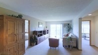 This single-floor space centers around a spacious living area finished with soft, wall-to-wall carpeting that lends a warm and inviting atmosphere throughout. The room is naturally illuminated by a large sliding glass door that opens to the outdoors, allowing ample daylight to fill the interior and creating a seamless connection to the outside. Neutral-toned walls complement the light-colored ceiling, which features a textured finish that adds subtle visual interest overhead.

The living area is well-proportioned, offering enough room to accommodate a comfortable seating arrangement, including a sofa and accent chair, alongside a glass-topped side table. A substantial storage unit with shelving and cabinetry lines one wall, providing both display and organizational functionality. Decorative plants are positioned throughout, adding a natural, lived-in quality to the space.

A doorway with warm wood trim is visible to one side, offering a glimpse into an adjacent bedroom, suggesting a practical and connected floor plan. The overall ambiance is calm and understated, with a layout designed to balance open living space with accessible storage and natural light.