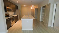 This single-floor residence offers a thoughtfully designed open-concept layout that seamlessly connects the kitchen and living areas. The kitchen serves as the heart of the space, anchored by a generously sized center island with a light stone countertop, an undermount sink, and elegant pendant lighting overhead. Dark espresso cabinetry lines the kitchen walls, providing a striking contrast to the light hardwood-style flooring that extends throughout the unit. Stainless steel appliances, including a refrigerator and range, complete the kitchen's polished, contemporary look. Track lighting along the kitchen ceiling adds additional illumination and a modern touch. The open layout flows naturally toward the main living area, creating a sense of spaciousness and ease of movement. A built-in shelving unit is conveniently positioned near the entryway, offering practical storage and display space. Neutral green-toned walls and warm, consistent flooring throughout lend the space a cohesive and inviting ambiance. The overall design balances functionality with modern aesthetic appeal, making this a well-appointed and comfortable single-level residence.