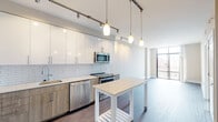 Located at 33 Monroe St in the heart of Rockville, MD, this modern single-floor residence offers a thoughtfully designed open-concept layout with clean lines and contemporary finishes throughout. The living area is bright and airy, anchored by expansive floor-to-ceiling windows that frame elevated views of the surrounding cityscape, flooding the space with natural light. Dark hardwood-style flooring runs throughout the main living areas, adding warmth and contrast to the neutral palette.

The kitchen is a standout feature, equipped with stainless steel appliances including a refrigerator, microwave, range, and dishwasher. Two-tone cabinetry pairs white upper cabinets with wood-finish lower cabinets, complemented by a classic white subway tile backsplash and a portable island with a light stone countertop. Pendant lighting and a track lighting system provide functional and stylish illumination.

The bedroom is generously proportioned with plush carpeting and a large window offering sweeping urban views and abundant natural light. The bathroom is well-appointed with light tile flooring, a full soaking tub with shower, a vanity with wood cabinetry, and clean white finishes. Multiple closets are accessible from the main hallway, providing ample storage throughout the residence.