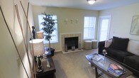 This single-floor living space offers a warm and inviting atmosphere, anchored by a centrally located tiled fireplace that serves as the room's focal point. The fireplace surround features clean, light-colored tile work with a simple white mantel, adding a classic touch to the overall design. The room is finished with wall-to-wall carpeting in a neutral beige tone, contributing to the cozy ambiance underfoot.

Three windows, dressed with horizontal blinds, allow generous natural light to fill the space, brightening the soft yellow-green painted walls and creating an airy feel throughout. A door with an adjacent window on one side suggests access to an outdoor area, further enhancing the sense of openness.

The living area accommodates a functional furniture arrangement, including a dark wood console table, a glass-top coffee table, and a sectional sofa. Decorative elements such as table lamps, lantern-style accessories, and a large indoor tree plant add character and a lived-in warmth to the space. Wall-mounted sconce fixtures provide additional ambient lighting options, rounding out this comfortable, well-appointed interior.
