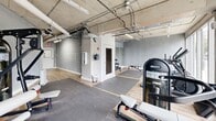 This fitness center in Miami's Brickell area offers a thoughtfully designed exercise space with an industrial-modern aesthetic. The room features exposed concrete ceilings with visible ductwork and piping, lending an urban, loft-style character throughout. Light wood-tone flooring contrasts with the raw concrete walls and gray painted surfaces, creating a clean and functional environment.

Large floor-to-ceiling windows and sliding glass doors line one side of the space, flooding the interior with natural light and providing street-level views of the surrounding neighborhood. The open layout accommodates a comprehensive selection of fitness equipment, including cardio machines such as treadmills, ellipticals, and a stationary bike, alongside a robust array of strength-training equipment including cable machines, Nautilus weight machines, a multi-station functional trainer, adjustable benches, and free weights. Protective rubber floor mats are positioned throughout the workout zones for safety and comfort.

A dedicated mirrored area supports stretching and floor exercises, while a small enclosed utility room provides additional infrastructure support. Strip lighting mounted to the ceiling ensures even, practical illumination across the entire floor. The space is well-suited for a full-body workout routine, offering both cardiovascular and resistance training options in a compact yet efficiently organized setting.