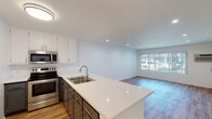 Located at 14960 Sherman Way in Van Nuys, California, this single-floor unit offers a clean, modern aesthetic with thoughtful updates throughout. The open-concept living and kitchen area is anchored by light wood-style flooring that extends seamlessly across the space, creating a bright and cohesive feel. Recessed lighting illuminates the interior, complemented by natural light streaming in through a large window that overlooks the exterior.

The kitchen is well-appointed with two-tone cabinetry — crisp white upper cabinets paired with darker lower cabinets — topped with quartz-style countertops. A peninsula with an extended breakfast bar provides additional prep space and casual seating. Stainless steel appliances, including an electric range and over-the-range microwave, complete the functional and stylish kitchen setup. The open sightline from the kitchen into the living area makes the floor plan feel spacious and connected.

The living area is generously sized, offering a versatile layout with a window unit for climate control and a large window dressed with vertical blinds, allowing for adjustable natural light. The bathroom features a full-size tub-shower combination with white surround, a toilet, and neutral wall tones. Throughout the unit, a consistent palette of whites and warm neutrals contributes to a clean, move-in-ready atmosphere.