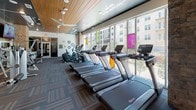 This fitness center is a comprehensively designed, single-floor facility that offers a diverse range of workout zones to support a full-body training experience. The space is characterized by a striking combination of warm wood-paneled ceilings, patterned carpet tiles, and polished hardwood flooring, creating a dynamic yet cohesive aesthetic throughout.

The cardio zone is a standout feature, lined with an extensive row of treadmills, ellipticals, and upright bikes positioned directly against floor-to-ceiling windows. These expansive windows flood the area with natural light and provide open views of the surrounding urban streetscape. Motivational phrases such as "Raise Your Heart Rate" and "Work Your Muscles" are displayed directly on the walls, reinforcing the space's energetic atmosphere.

A dedicated cycling and yoga studio offers a bright, open area with hardwood floors, spin bikes, purple yoga mats, and a large wall-mounted TV. Floor-to-ceiling mirrored glass panels visually expand the room while connecting it to the broader fitness floor.

A functional training zone features a turf-accent wall, TRX suspension systems, medicine balls, and resistance equipment, ideal for high-intensity interval and strength training. Exposed brick columns add an industrial design element, while ceiling fans and recessed lighting ensure comfort throughout the entire facility.