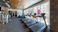 This fitness center is a comprehensively designed, single-floor facility that offers a diverse range of workout zones to support a full-body training experience. The space is characterized by a striking combination of warm wood-paneled ceilings, patterned carpet tiles, and polished hardwood flooring, creating a dynamic yet cohesive aesthetic throughout.

The cardio zone is a standout feature, lined with an extensive row of treadmills, ellipticals, and upright bikes positioned directly against floor-to-ceiling windows. These expansive windows flood the area with natural light and provide open views of the surrounding urban streetscape. Motivational phrases such as "Raise Your Heart Rate" and "Work Your Muscles" are displayed directly on the walls, reinforcing the space's energetic atmosphere.

A dedicated cycling and yoga studio offers a bright, open area with hardwood floors, spin bikes, purple yoga mats, and a large wall-mounted TV. Floor-to-ceiling mirrored glass panels visually expand the room while connecting it to the broader fitness floor.

A functional training zone features a turf-accent wall, TRX suspension systems, medicine balls, and resistance equipment, ideal for high-intensity interval and strength training. Exposed brick columns add an industrial design element, while ceiling fans and recessed lighting ensure comfort throughout the entire facility.