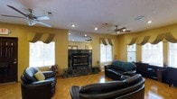This single-floor space exudes a comfortable and welcoming ambiance, thoughtfully designed for relaxed living and entertaining. The centerpiece of the main living area is a stunning floor-to-ceiling marble fireplace finished in deep green and black tones, adding a bold and elegant focal point to the room. Surrounding the fireplace, warm yellow-toned walls create an inviting atmosphere that complements the rich hardwood parquet flooring below. The living area is generously sized, comfortably accommodating a full suite of leather seating, and benefits from an abundance of natural light streaming through multiple large windows dressed with layered curtains and horizontal blinds. Ceiling fans are installed throughout, providing both functional air circulation and a classic decorative touch. Recessed lighting supplements the natural light, ensuring the space is well-lit at all times. The textured ceiling adds subtle visual depth, while the overall layout flows naturally, offering a sense of openness across the single floor. The combination of rich materials, warm colors, and thoughtful design details makes this a space that is both functional and full of character.