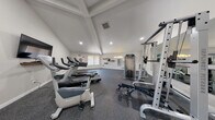 This single-floor fitness facility is thoughtfully designed to support a full range of workout needs in a clean, welcoming environment. The space features vaulted ceilings with exposed beam detailing and recessed lighting, lending an open and airy atmosphere throughout. Durable speckled rubber flooring covers the entire workout area, providing both safety and functionality underfoot.

The cardio section is well-appointed with multiple treadmills and stationary bikes, complemented by a wall-mounted flat-screen television for entertainment during workouts. Strength training is thoroughly covered with a multi-station cable machine, a power rack, a weight bench, and a full rack of dumbbells in graduated sizes. A large wall mirror enhances the sense of space while allowing users to monitor their form during exercises.

A dedicated stretching and flexibility zone features wall-mounted wooden shelves holding yoga mats and accessories, wall-mounted stability balls, and resistance bands neatly hung for easy access. A motivational neon sign reading "you got this" adds an energetic, personalized touch to the space. Natural light filters in through a window with vertical blinds, and the neutral white walls keep the environment feeling clean and focused. The facility also includes a security camera and clearly posted safety signage, reflecting a commitment to user well-being.