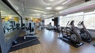 This fitness center offers a spacious, single-floor layout designed to accommodate a comprehensive workout experience. The room features warm hardwood flooring throughout, providing a clean and professional aesthetic that pairs well with the bright, airy atmosphere created by a generous row of large windows along one wall. These expansive windows invite abundant natural light while offering views of the surrounding outdoor landscape, enhancing the overall workout environment.

The ceiling is a standout architectural feature, finished in white with exposed structural elements and accented by large, circular pendant light fixtures that contribute to the modern, stylish ambiance. Track lighting supplements the natural light, ensuring the space is well-lit at all times.

The fitness floor is thoughtfully organized to accommodate both cardio and strength training. Cardio equipment includes multiple treadmills and elliptical machines, each equipped with individual screens for entertainment during workouts. The strength training area features cable machines, weight benches, and various resistance equipment, all arranged to allow for comfortable, unobstructed movement between stations. Full-length mirrors on one wall support form correction and create a sense of expanded space.

Adding a creative touch, a vibrant, colorful mural adorns one wall, infusing the space with energy and character. The overall design balances functionality with an inviting atmosphere, making it a well-rounded fitness facility.